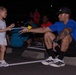 A young girl with face paint and colorful stickers on her leg is handing or receiving a glow stick from a tattooed adult male crouching on a parking lot curb stop at night. The scene appears to be an outdoor nighttime event, likely a fun run or glow-themed community gathering. Several other participants are visible in the background, some wearing glow accessories.