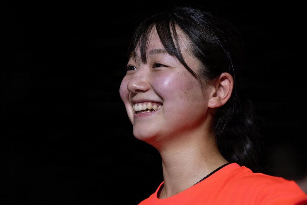 A young woman is photographed in a three-quarter profile view against a dark background, displaying a broad, genuine smile. She is wearing a bright orange/red top and has dark hair pulled back. The lighting highlights her face dramatically against the black background.