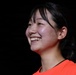 A young woman is photographed in a three-quarter profile view against a dark background, displaying a broad, genuine smile. She is wearing a bright orange/red top and has dark hair pulled back. The lighting highlights her face dramatically against the black background.