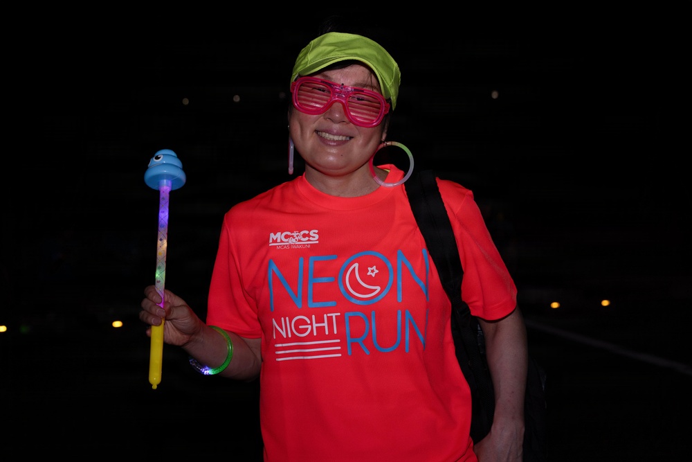 A smiling woman poses at night during a 'Neon Night Run' event organized by MCCS MCAS Iwakuni. She is dressed in festive, colorful accessories including novelty shutter shades, a neon green cap, and large hoop earrings, and is holding a light-up toy wand. The dark background and glowing accessories are consistent with a nighttime fun run event.