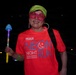A smiling woman poses at night during a 'Neon Night Run' event organized by MCCS MCAS Iwakuni. She is dressed in festive, colorful accessories including novelty shutter shades, a neon green cap, and large hoop earrings, and is holding a light-up toy wand. The dark background and glowing accessories are consistent with a nighttime fun run event.