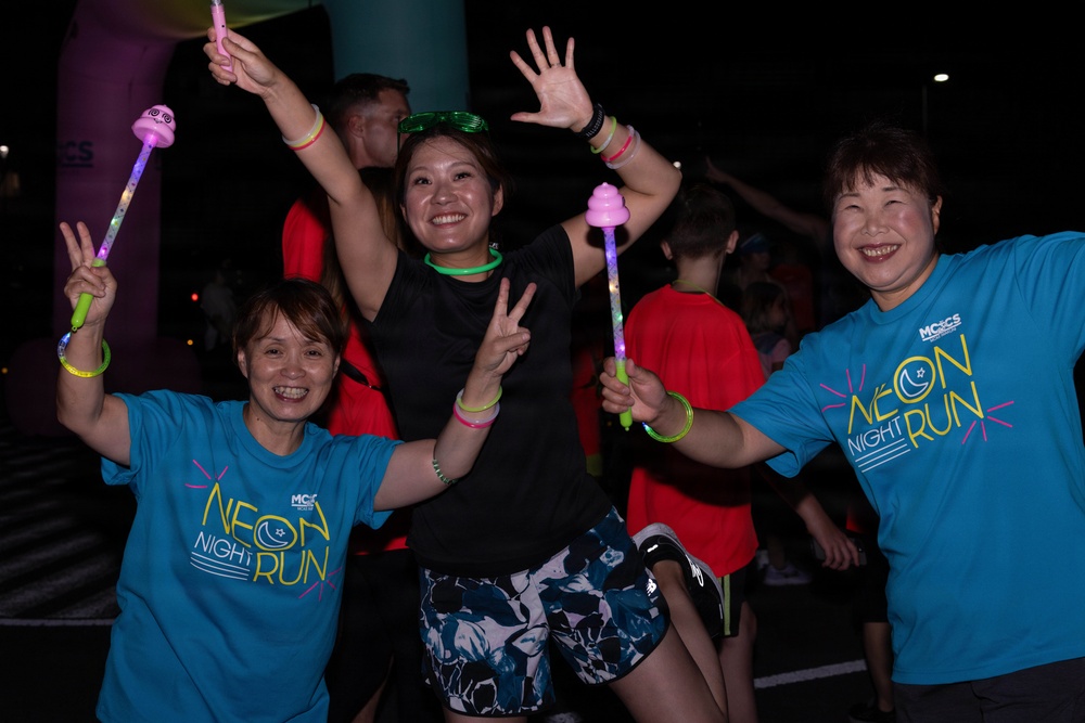 Three women pose enthusiastically at a Neon Night Run event, likely organized by MCCS (Marine Corps Community Services). Two of the women wear matching teal 'Neon Night Run' t-shirts, while the center woman wears a black athletic outfit with neon accessories. They are holding illuminated light-up wands and making celebratory gestures at what appears to be a nighttime fun run event.