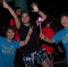 Three women pose enthusiastically at a Neon Night Run event, likely organized by MCCS (Marine Corps Community Services). Two of the women wear matching teal 'Neon Night Run' t-shirts, while the center woman wears a black athletic outfit with neon accessories. They are holding illuminated light-up wands and making celebratory gestures at what appears to be a nighttime fun run event.