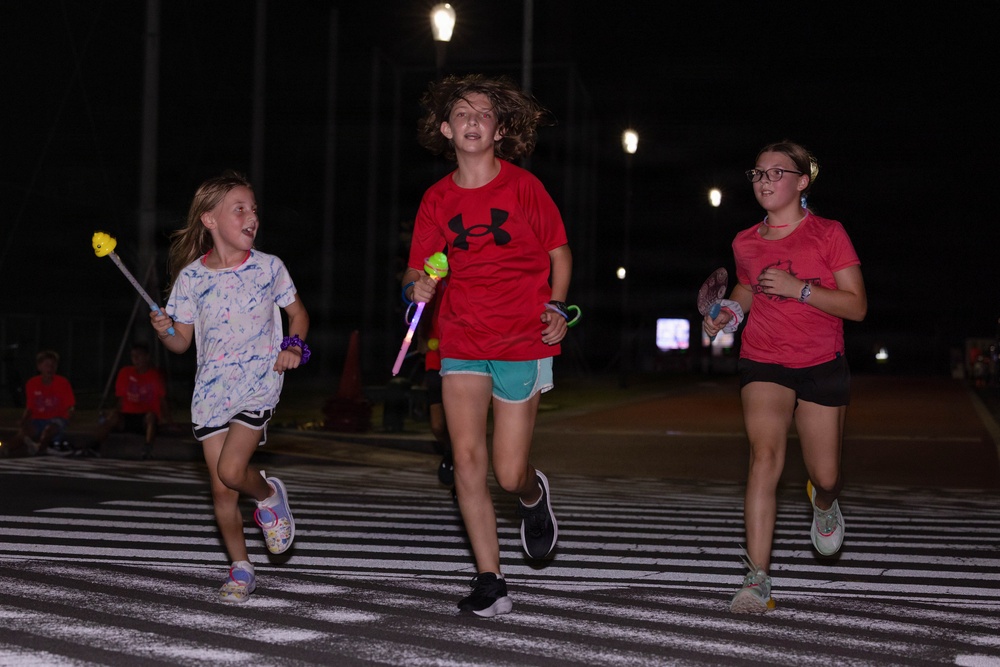 Three children are running across a crosswalk at night, appearing to participate in a fun run or nighttime event. The middle child wears a red Under Armour shirt and carries a glowing toy, while the other two children also hold light-up or novelty items. Street lights illuminate the scene against a dark background.