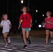 Three children are running across a crosswalk at night, appearing to participate in a fun run or nighttime event. The middle child wears a red Under Armour shirt and carries a glowing toy, while the other two children also hold light-up or novelty items. Street lights illuminate the scene against a dark background.