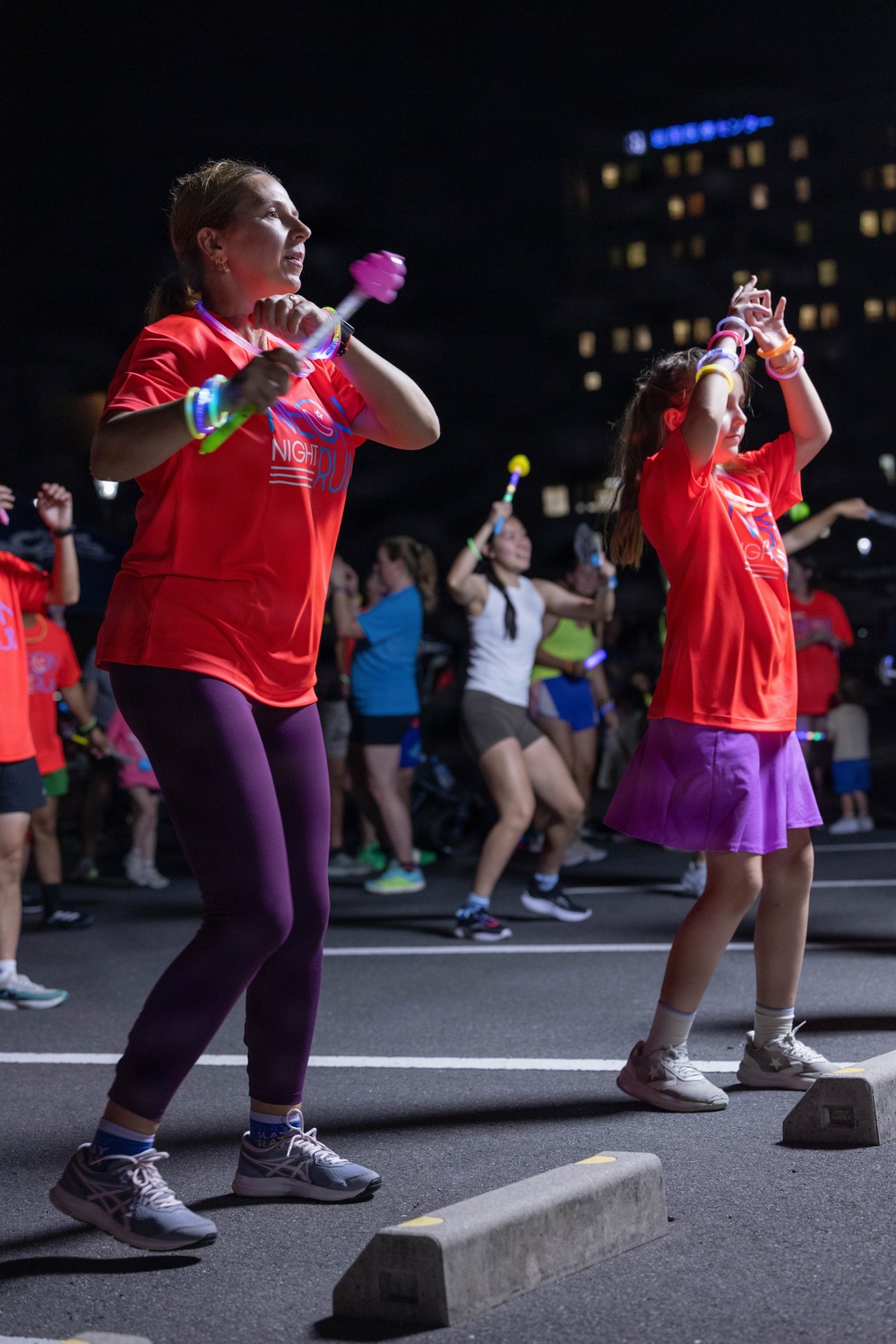 A nighttime outdoor event featuring a large group of people participating in a dance or exercise activity. Two individuals in the foreground wear matching red 'Night Run' t-shirts and hold glowing light-up accessories, with a crowd of similarly dressed participants visible behind them. The event takes place on a street or parking area with an illuminated city building visible in the background.