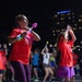 A nighttime outdoor event featuring a large group of people participating in a dance or exercise activity. Two individuals in the foreground wear matching red 'Night Run' t-shirts and hold glowing light-up accessories, with a crowd of similarly dressed participants visible behind them. The event takes place on a street or parking area with an illuminated city building visible in the background.