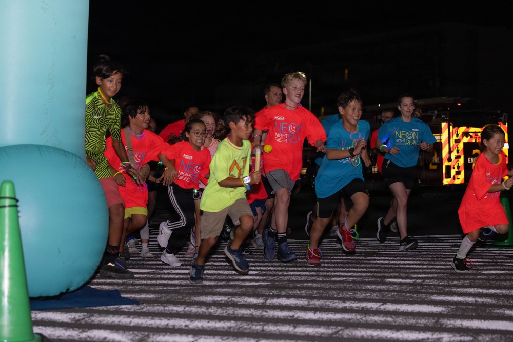 A group of children are participating in a 'Neon Night Run' event, captured at the starting line during nighttime. Many participants are wearing bright neon-colored shirts (red, teal, yellow-green) branded with 'Neon Night Run' logos, along with glow accessories. The event appears to be an organized youth fun run with inflatable decorations and illuminated signage in the background.