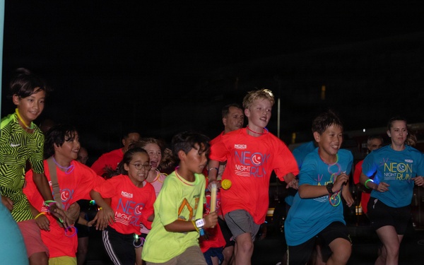 A group of children are participating in a 'Neon Night Run' event, captured at the starting line during nighttime. Many participants are wearing bright neon-colored shirts (red, teal, yellow-green) branded with 'Neon Night Run' logos, along with glow accessories. The event appears to be an organized youth fun run with inflatable decorations and illuminated signage in the background.