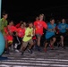 A group of children are participating in a 'Neon Night Run' event, captured at the starting line during nighttime. Many participants are wearing bright neon-colored shirts (red, teal, yellow-green) branded with 'Neon Night Run' logos, along with glow accessories. The event appears to be an organized youth fun run with inflatable decorations and illuminated signage in the background.
