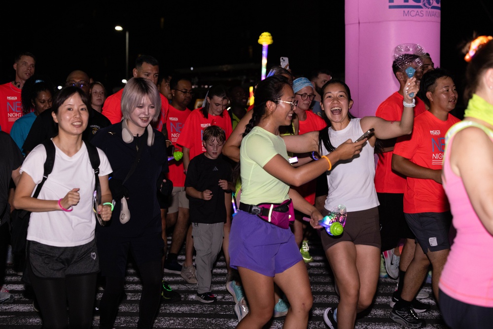 A nighttime fun run or glow run event is taking place, with participants wearing red event t-shirts and glow accessories. The crowd appears energetic and celebratory, with several participants smiling, laughing, and high-fiving near what appears to be a start or finish line marked by a pink inflatable arch. Partial text on shirts and the arch suggests this is an MCAS Iwakuni (Marine Corps Air Station) NEX Night Run event.