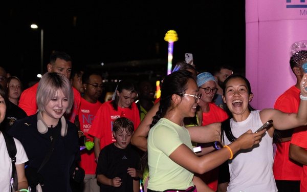 A nighttime fun run or glow run event is taking place, with participants wearing red event t-shirts and glow accessories. The crowd appears energetic and celebratory, with several participants smiling, laughing, and high-fiving near what appears to be a start or finish line marked by a pink inflatable arch. Partial text on shirts and the arch suggests this is an MCAS Iwakuni (Marine Corps Air Station) NEX Night Run event.