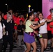 A nighttime fun run or glow run event is taking place, with participants wearing red event t-shirts and glow accessories. The crowd appears energetic and celebratory, with several participants smiling, laughing, and high-fiving near what appears to be a start or finish line marked by a pink inflatable arch. Partial text on shirts and the arch suggests this is an MCAS Iwakuni (Marine Corps Air Station) NEX Night Run event.