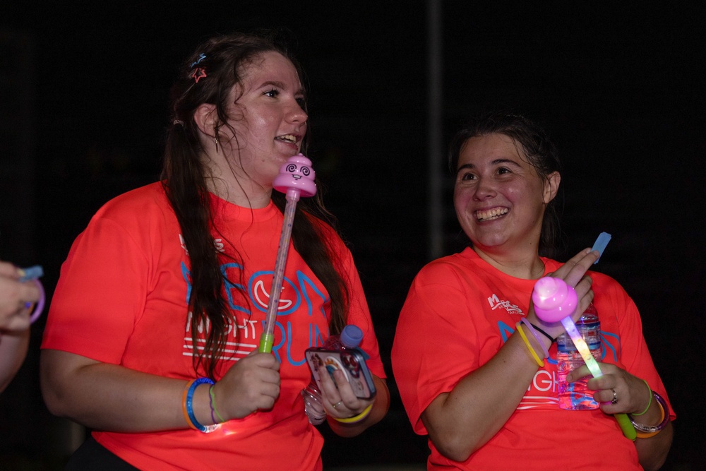 Two young women wearing matching orange/coral t-shirts with 'Neon Night Run' text are photographed at night during what appears to be a fun run event. Both are smiling and holding illuminated glow toys/wands, and are wearing glow bracelets. The image is taken in low-light nighttime conditions with the subjects well-lit by a flash or nearby light source.