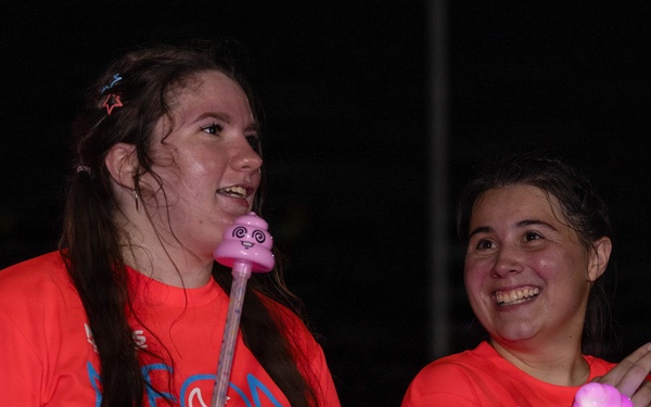 Two young women wearing matching orange/coral t-shirts with 'Neon Night Run' text are photographed at night during what appears to be a fun run event. Both are smiling and holding illuminated glow toys/wands, and are wearing glow bracelets. The image is taken in low-light nighttime conditions with the subjects well-lit by a flash or nearby light source.