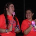 Two young women wearing matching orange/coral t-shirts with 'Neon Night Run' text are photographed at night during what appears to be a fun run event. Both are smiling and holding illuminated glow toys/wands, and are wearing glow bracelets. The image is taken in low-light nighttime conditions with the subjects well-lit by a flash or nearby light source.