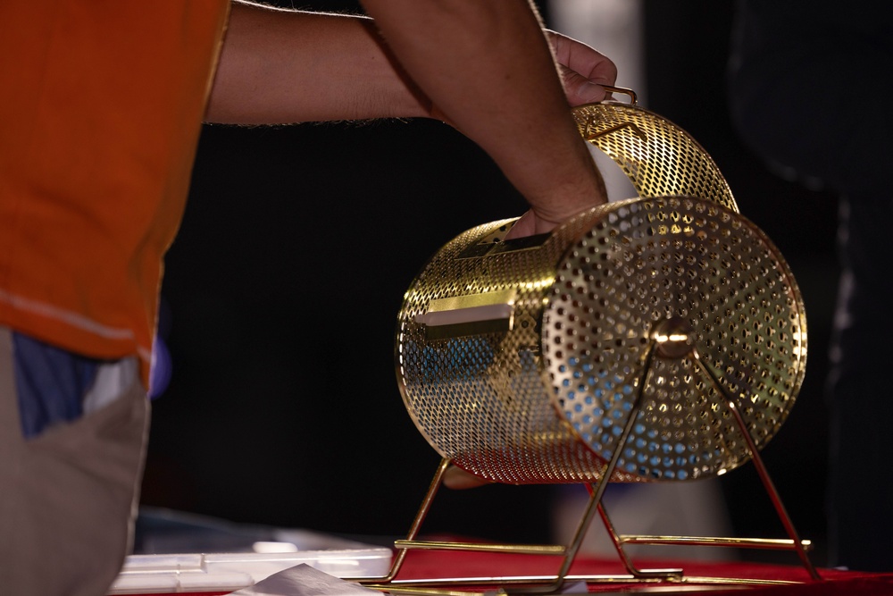 A person wearing an orange shirt is handling a gold-colored rotating lottery or raffle drum cage mounted on a stand. The cylindrical mesh drum appears to be a classic bingo or raffle ticket tumbler made of perforated gold-toned metal. The device is positioned on a red surface with a dark background.