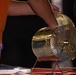 A person wearing an orange shirt is handling a gold-colored rotating lottery or raffle drum cage mounted on a stand. The cylindrical mesh drum appears to be a classic bingo or raffle ticket tumbler made of perforated gold-toned metal. The device is positioned on a red surface with a dark background.
