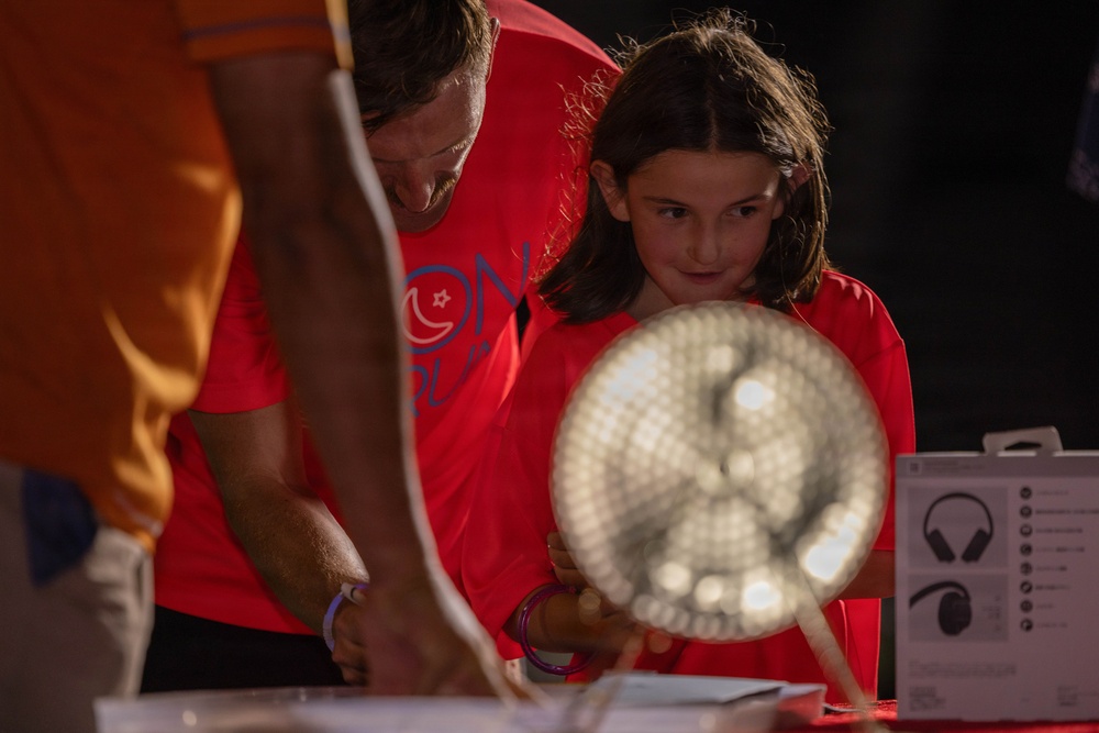 A child and an adult, both wearing red t-shirts, are engaged at what appears to be an interactive exhibit or activity table. The child is holding a circular illuminated object, possibly a light-up fan or display device, and appears focused and curious. A third person in an orange shirt is partially visible in the foreground.
