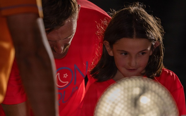 A child and an adult, both wearing red t-shirts, are engaged at what appears to be an interactive exhibit or activity table. The child is holding a circular illuminated object, possibly a light-up fan or display device, and appears focused and curious. A third person in an orange shirt is partially visible in the foreground.