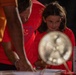 A child and an adult, both wearing red t-shirts, are engaged at what appears to be an interactive exhibit or activity table. The child is holding a circular illuminated object, possibly a light-up fan or display device, and appears focused and curious. A third person in an orange shirt is partially visible in the foreground.