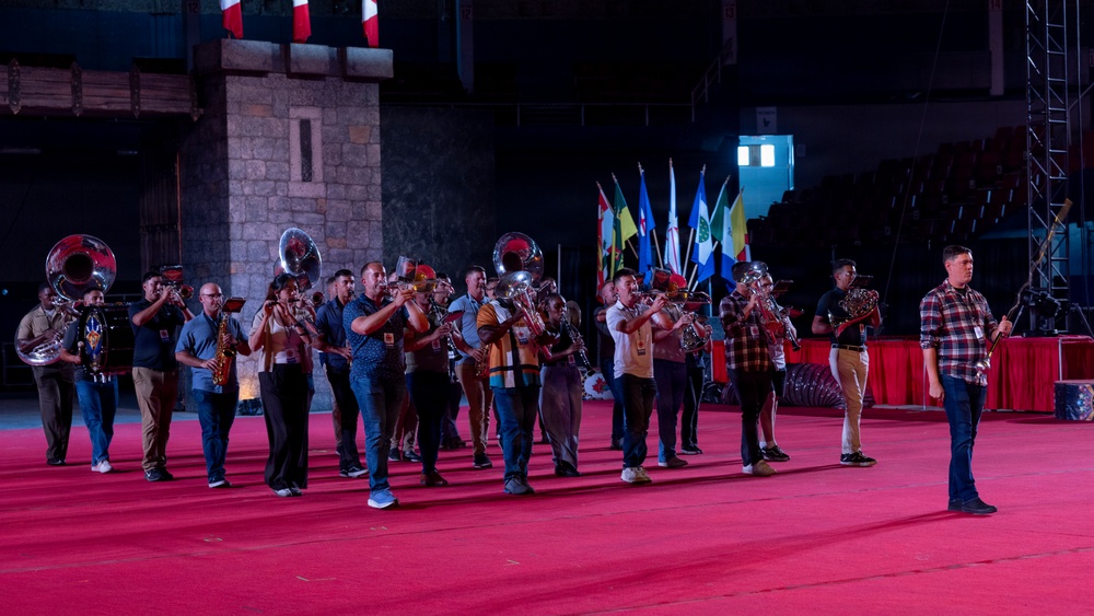 A marching band rehearsal or performance is taking place on a large red-carpeted floor inside what appears to be an indoor arena or event venue. The musicians are playing brass and wind instruments while a conductor or drum major stands apart to the right, facing the group. Multiple international flags are visible in the background, suggesting an international festival or ceremonial event.