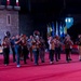 A marching band rehearsal or performance is taking place on a large red-carpeted floor inside what appears to be an indoor arena or event venue. The musicians are playing brass and wind instruments while a conductor or drum major stands apart to the right, facing the group. Multiple international flags are visible in the background, suggesting an international festival or ceremonial event.