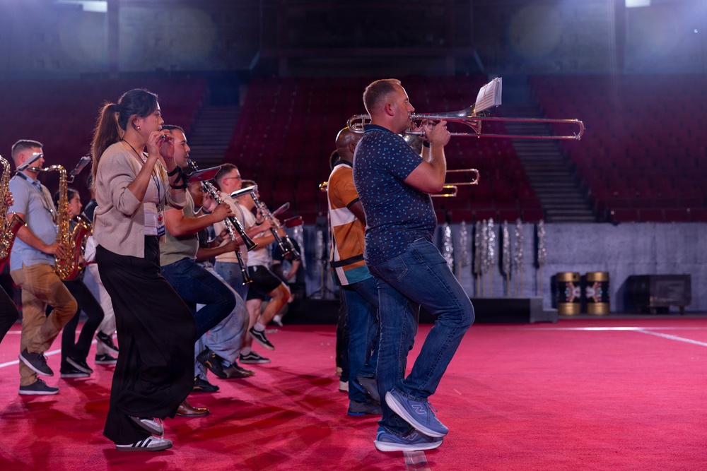 A group of musicians are rehearsing or performing inside a large indoor arena or stadium with red seating visible in the background. The ensemble includes players of various wind instruments including trombone, clarinet, and saxophone. The performers are on a red floor surface, dressed in casual clothing, suggesting a rehearsal rather than a formal performance.