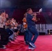 A group of musicians are rehearsing or performing inside a large indoor arena or stadium with red seating visible in the background. The ensemble includes players of various wind instruments including trombone, clarinet, and saxophone. The performers are on a red floor surface, dressed in casual clothing, suggesting a rehearsal rather than a formal performance.