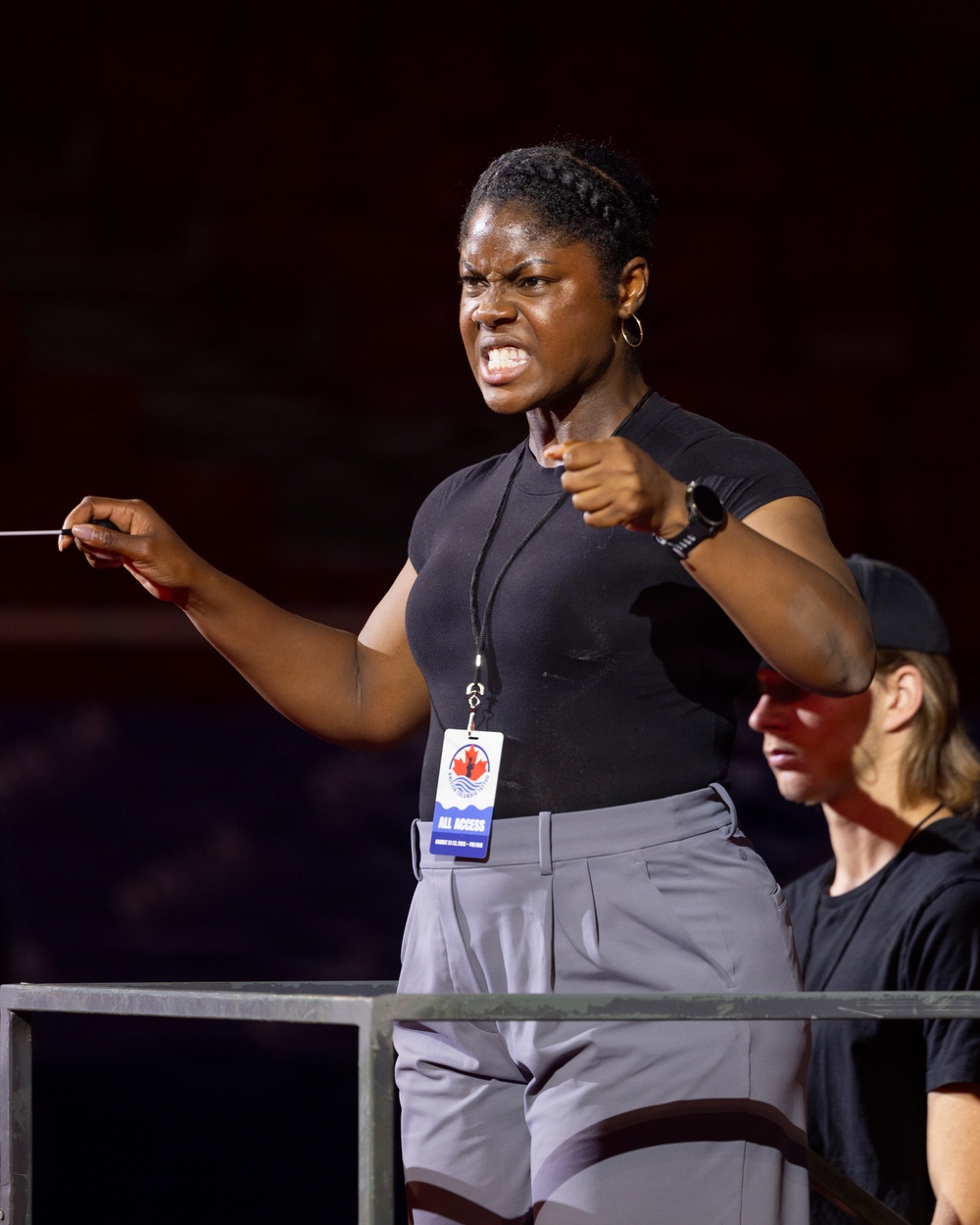 A woman with an intense, focused expression is conducting or directing, holding a baton in her left hand while pointing with her right fist. She wears a black t-shirt, gray pants, a watch, hoop earrings, and an 'All Access' credential badge featuring a Canadian maple leaf logo. A second person wearing a black cap and sunglasses is partially visible in the background.