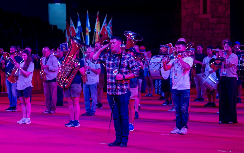 A nighttime outdoor band performance or tattoo event featuring a large ensemble of musicians playing brass and percussion instruments under dramatic pink/purple stage lighting. Multiple flags are visible in the background, suggesting a ceremonial or national event. A central figure in a plaid shirt appears to be conducting or directing the ensemble while saluting.