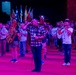 A nighttime outdoor band performance or tattoo event featuring a large ensemble of musicians playing brass and percussion instruments under dramatic pink/purple stage lighting. Multiple flags are visible in the background, suggesting a ceremonial or national event. A central figure in a plaid shirt appears to be conducting or directing the ensemble while saluting.
