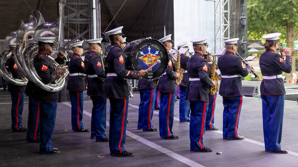 A U.S. Marine Corps military band is performing on an outdoor stage. The band is identified as the 'Third Marine Aircraft Wing Band' as visible on the large bass drum. Musicians are dressed in formal dress blue uniforms with white covers (caps) and are playing various instruments.