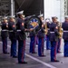 A U.S. Marine Corps military band is performing on an outdoor stage. The band is identified as the 'Third Marine Aircraft Wing Band' as visible on the large bass drum. Musicians are dressed in formal dress blue uniforms with white covers (caps) and are playing various instruments.