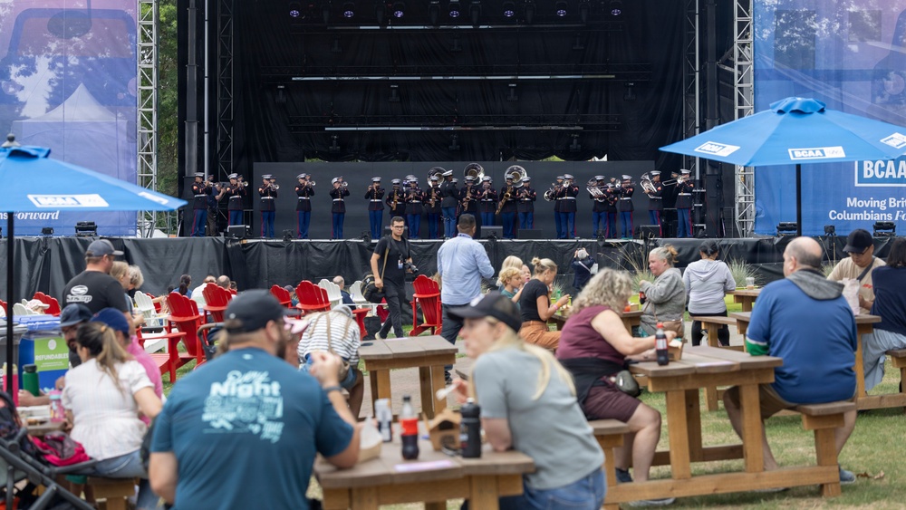 An outdoor public event featuring a military marching band performing on a large stage while audience members dine at wooden picnic tables and red Adirondack chairs in the foreground. The performers appear to be U.S. Marines in dress uniforms playing brass instruments. Blue BCAA-branded umbrellas provide shade for attendees at what appears to be a festival or community event.