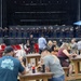 An outdoor public event featuring a military marching band performing on a large stage while audience members dine at wooden picnic tables and red Adirondack chairs in the foreground. The performers appear to be U.S. Marines in dress uniforms playing brass instruments. Blue BCAA-branded umbrellas provide shade for attendees at what appears to be a festival or community event.