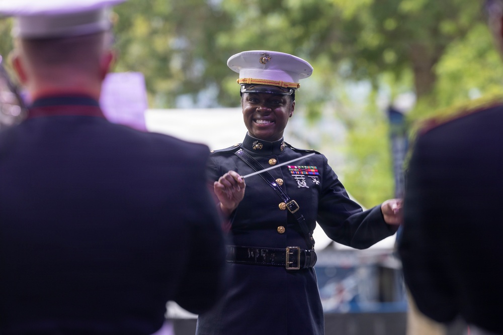 A U.S. Marine Corps officer in dress blues uniform is conducting a military band outdoors, holding a conductor's baton and smiling. She is positioned facing other uniformed Marines who are visible in the foreground as blurred figures. The setting appears to be an outdoor public performance or ceremony.