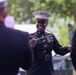 A U.S. Marine Corps officer in dress blues uniform is conducting a military band outdoors, holding a conductor's baton and smiling. She is positioned facing other uniformed Marines who are visible in the foreground as blurred figures. The setting appears to be an outdoor public performance or ceremony.