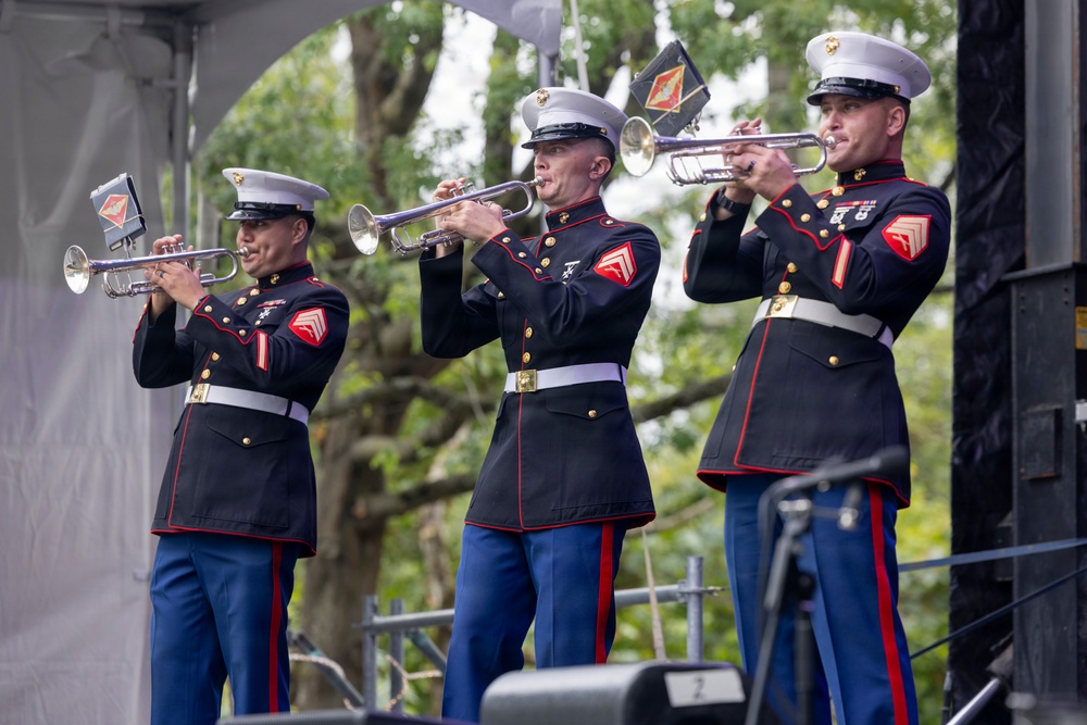 Three United States Marine Corps musicians in dress blues are performing on an outdoor stage, each playing a trumpet or bugle. They are wearing the distinctive USMC dress uniform with white covers (caps), blue trousers with red stripes, and gold buttons. The setting appears to be a formal public ceremony or military performance event.