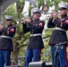 Three United States Marine Corps musicians in dress blues are performing on an outdoor stage, each playing a trumpet or bugle. They are wearing the distinctive USMC dress uniform with white covers (caps), blue trousers with red stripes, and gold buttons. The setting appears to be a formal public ceremony or military performance event.