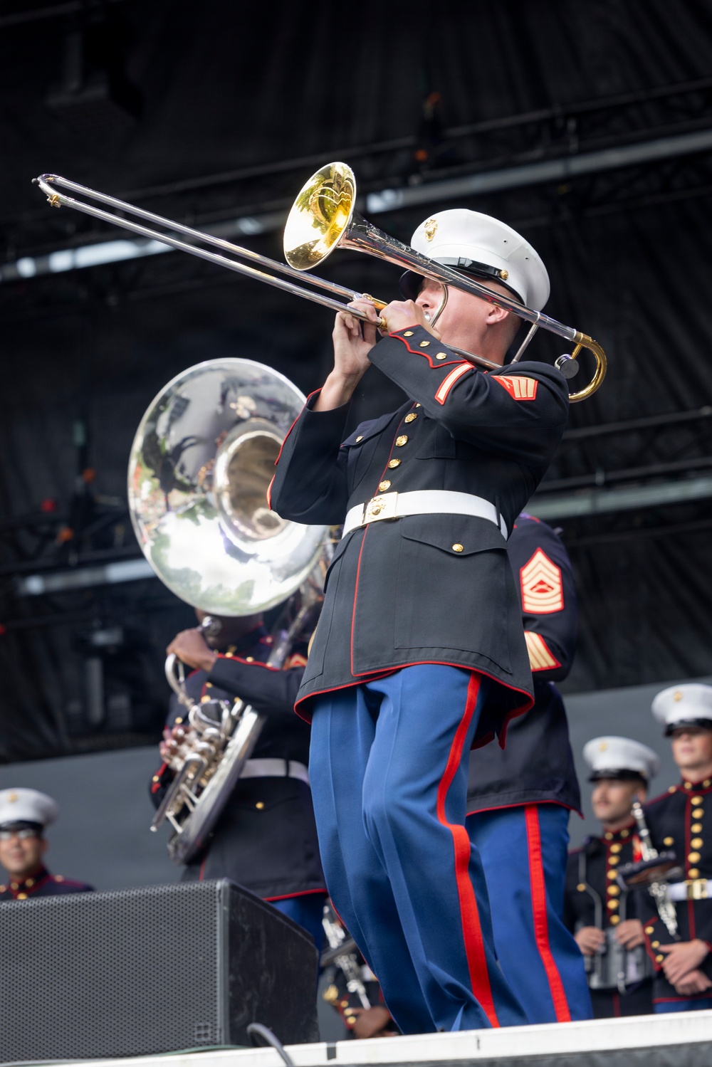 A United States Marine Corps band is performing on an outdoor stage. The central figure is a Marine playing a trombone while wearing the distinctive dress blues uniform with white cover, white belt, blue trousers with red stripe, and gold buttons. Additional band members are visible in the background playing a sousaphone and other instruments.