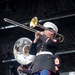 A United States Marine Corps band is performing on an outdoor stage. The central figure is a Marine playing a trombone while wearing the distinctive dress blues uniform with white cover, white belt, blue trousers with red stripe, and gold buttons. Additional band members are visible in the background playing a sousaphone and other instruments.