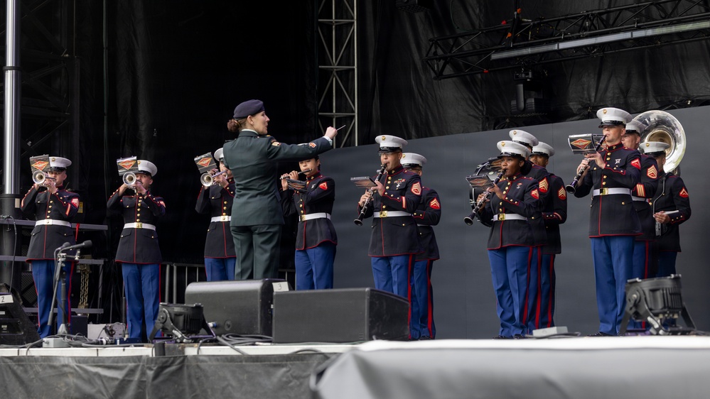 A military band, identifiable as U.S. Marines based on their distinctive dress blue uniforms with red stripes, performs on an outdoor stage. A conductor in a green uniform stands center-stage with baton raised, directing the ensemble. The band includes musicians playing clarinets, trumpets, flutes, and a sousaphone.