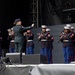 A military band, identifiable as U.S. Marines based on their distinctive dress blue uniforms with red stripes, performs on an outdoor stage. A conductor in a green uniform stands center-stage with baton raised, directing the ensemble. The band includes musicians playing clarinets, trumpets, flutes, and a sousaphone.