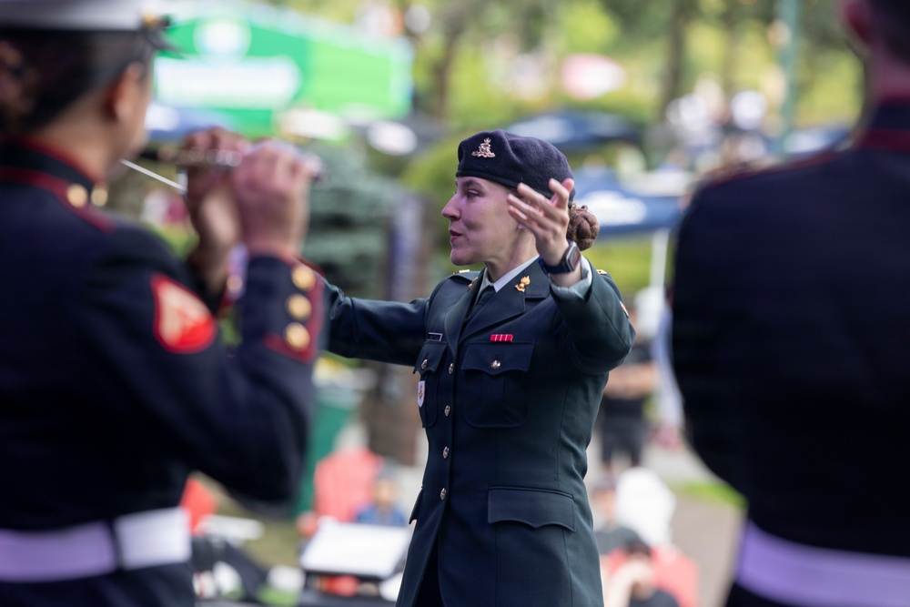 A female military officer in a dark green dress uniform and black beret is conducting a military band outdoors. She is gesturing with both hands toward musicians visible in the foreground, who are wearing dark uniforms with red trim. The setting appears to be a public outdoor event or ceremony.