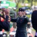 A female military officer in a dark green dress uniform and black beret is conducting a military band outdoors. She is gesturing with both hands toward musicians visible in the foreground, who are wearing dark uniforms with red trim. The setting appears to be a public outdoor event or ceremony.