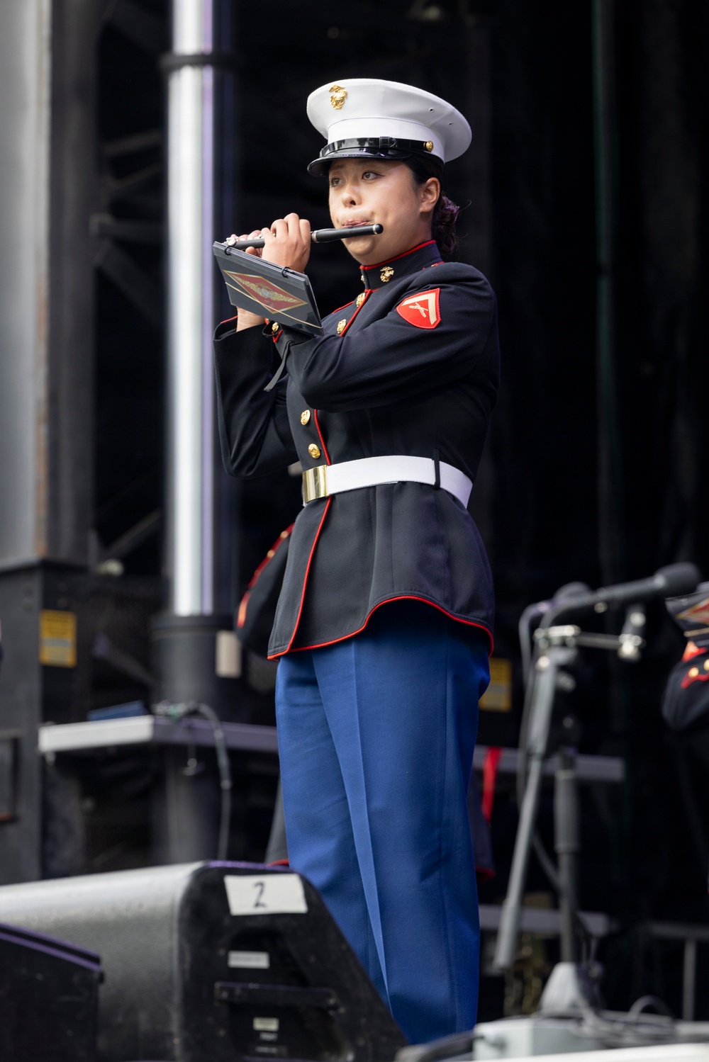 A United States Marine Corps musician in dress blues uniform is playing a flute on a stage. She is holding sheet music in a black folder while performing, suggesting a formal military band performance. The setting appears to be an outdoor stage with audio equipment visible in the background.