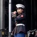 A United States Marine Corps musician in dress blues uniform is playing a flute on a stage. She is holding sheet music in a black folder while performing, suggesting a formal military band performance. The setting appears to be an outdoor stage with audio equipment visible in the background.