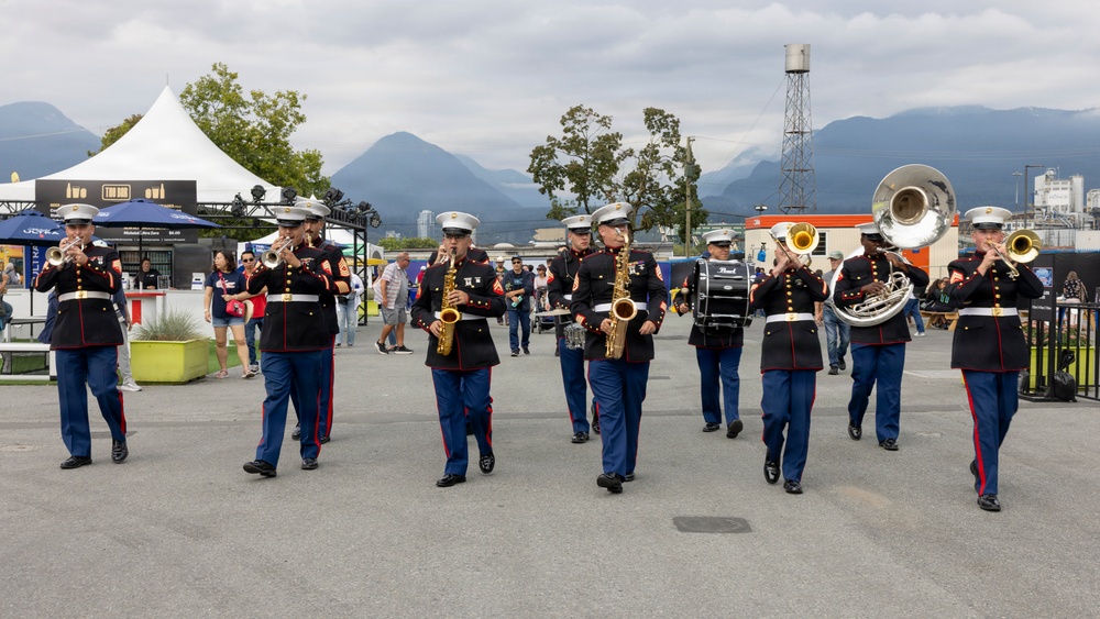 A United States Marine Corps marching band performs outdoors at what appears to be a public event or festival. The musicians are dressed in formal dress blues uniforms with white caps and are playing various instruments while marching across a paved plaza. Mountains and overcast skies are visible in the background, suggesting a Pacific Northwest location.