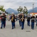 A United States Marine Corps marching band performs outdoors at what appears to be a public event or festival. The musicians are dressed in formal dress blues uniforms with white caps and are playing various instruments while marching across a paved plaza. Mountains and overcast skies are visible in the background, suggesting a Pacific Northwest location.